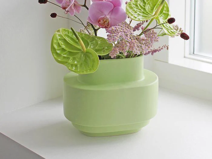 En lysegrøn vase med rosa og grønne blomster står i en hvid vindueskarm ved siden af et vindue med naturligt lysindfald.