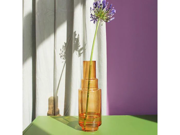 En enkelt lilla blomst i en ravfarvet glasvase står på en grøn flade, mens sollyset kaster skygger på et gardin og en lavendelfarvet væg i baggrunden.