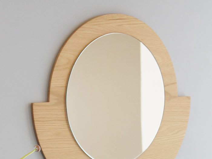 Ovalt spejl med lys træramme monteret på en lysegrå væg, der reflekterer en del af væggen og lyset.