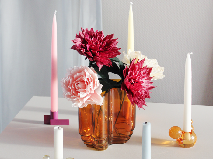 Ravfarvet glasvase med rosa, røde og cremefarvede blomster omgivet af høje lys i pastelfarver på en hvid overflade.