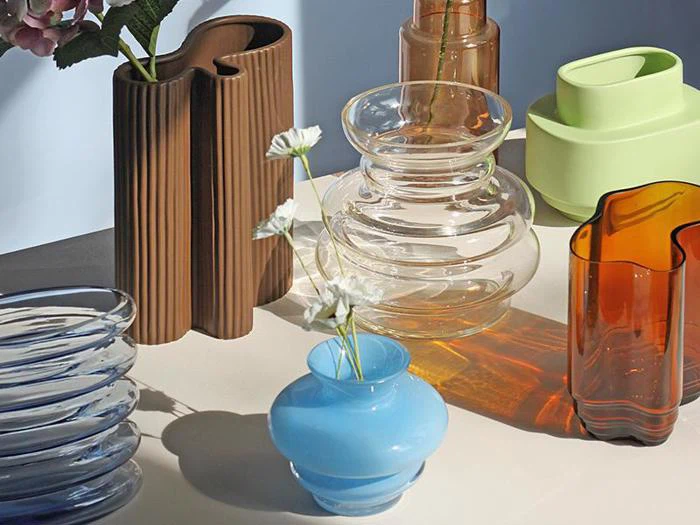 Forskellige farverige glas- og keramikvaser, bl.a. blå, ravfarvede, klare, brune og grønne, udstillet på en neutral overflade med nogle hvide blomster i en blå vase.