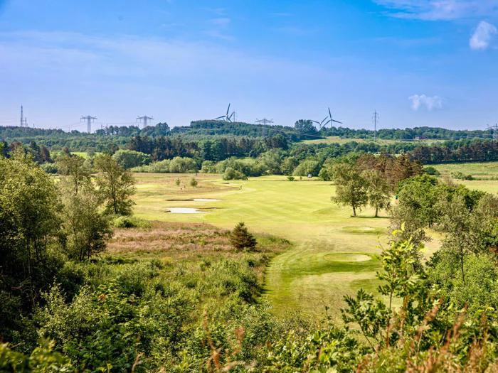 En frodig grøn golfbane med sandbunkers, omgivet af træer og marker; vindmøller og højspændingsledninger kan ses i den fjerne horisont under en blå himmel.