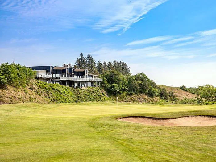 En moderne bygning har udsigt til en golfbane med en sandbunker, frodigt grønt græs og træer under en blå himmel med spredte skyer.