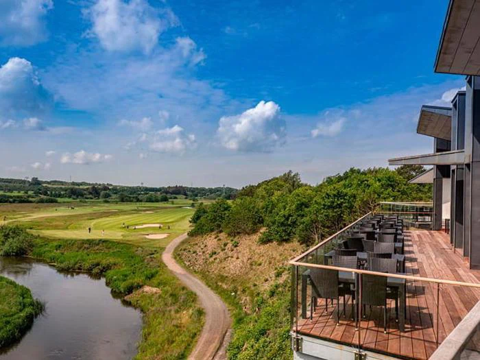 Fra en træterrasse med borde og stole er der udsigt over en golfbane, grønne marker og en snoet sti langs et vandløb under en blå himmel med spredte skyer.