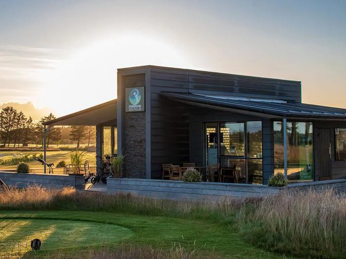 En moderne bygning med store vinduer ud til en golfbane ved solnedgang, med udendørs siddepladser, golfbags og græsklædte omgivelser.