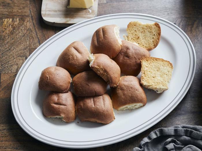 En hvid, oval tallerken med flere bagte, brune rundstykker, nogle hele og andre flækkede, på et træbord.