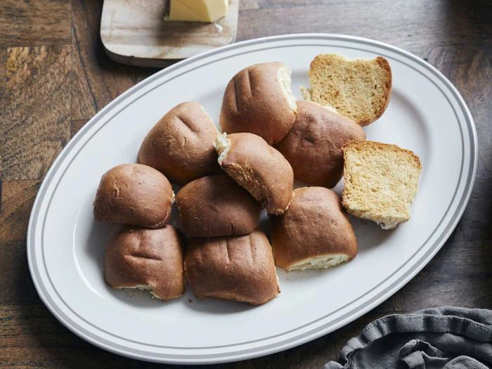 En hvid oval tallerken med flere brune rundstykker, nogle hele og andre flækkede, på et træbord.