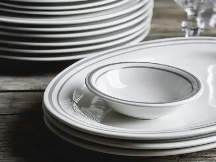 En stak hvide tallerkener og en lille skål med tynde grå kanter står på et træbord ved siden af glas.