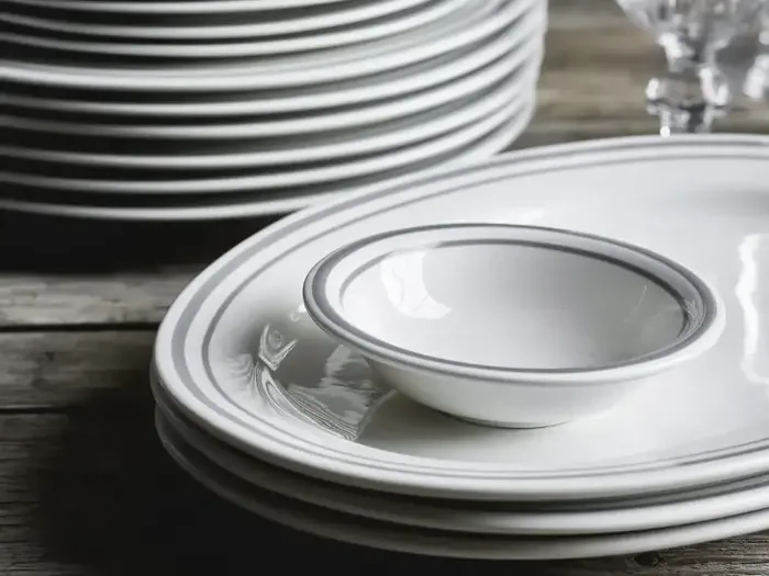 En stak hvide tallerkener med grå kant, en lille skål på toppen og klare glasvarer i baggrunden, placeret på en træoverflade.