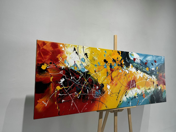 Et farverigt abstrakt maleri med dristige røde, gule, sorte, hvide og blå stænk og linjer, udstillet på et staffeli af træ mod en almindelig hvid væg.