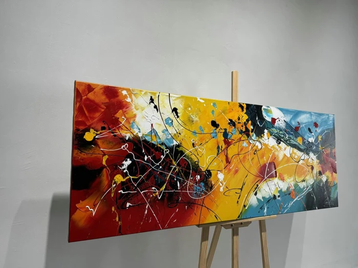 Et farverigt abstrakt maleri med dristige røde, gule, sorte, hvide og blå stænk og linjer, udstillet på et staffeli af træ mod en almindelig hvid væg.