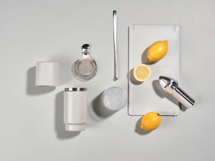 Et cocktailshaker-sæt, citroner, en halv citron og en citruspresser på en hvid flade med et lille skærebræt.