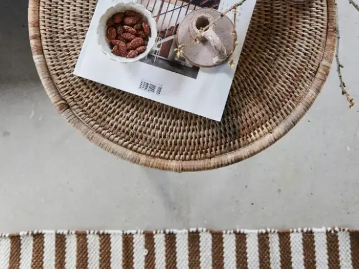 På et rundt flettet bord ligger et magasin, en lille skål med mandler og en beige keramikkop; en del af et stribet tæppe er synligt nedenunder.