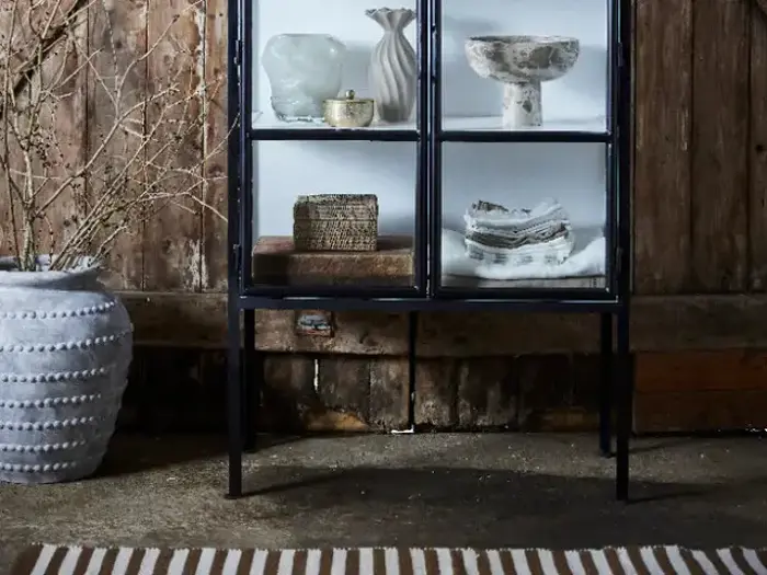Et vitrineskab med dekorative genstande ved siden af en stor struktureret hvid vase med nøgne grene, opstillet mod en rustik trævæg.