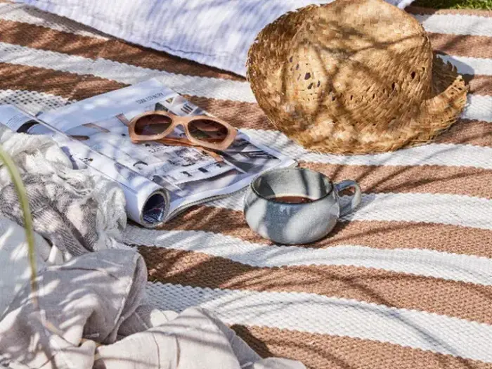 Et stribet picnictæppe med en stråhat, et sammenrullet magasin, solbriller, en kop kaffe og et let tørklæde, alt sammen badet i sollys og skygger.