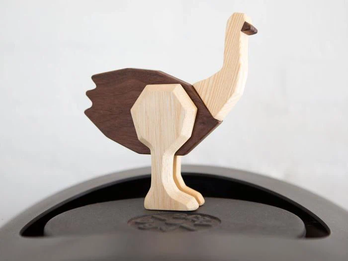 En strudsefigur af træ med lyse og mørke træpartier står på en glat, sort overflade mod en almindelig hvid baggrund.