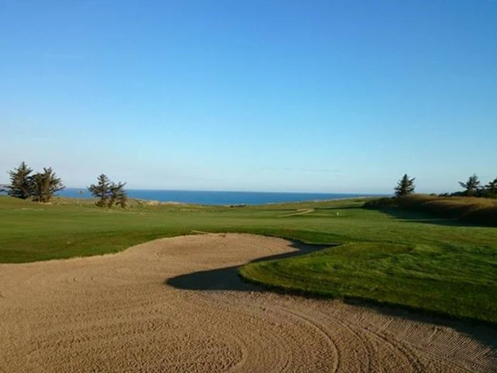 En golfbane med en sandbunker i forgrunden, grønne fairways, træer og en fjern udsigt over havet under en klar blå himmel.