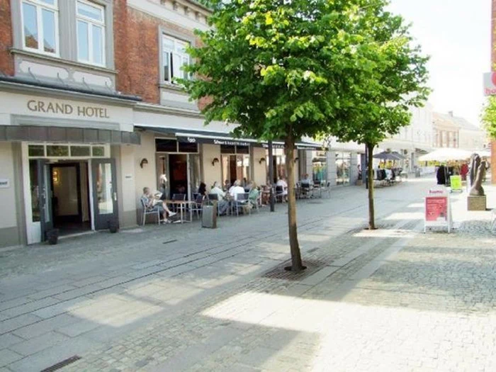 En solrig gade med udeservering på en café nær Grand Hotel, omkranset af små træer og folk, der spiser ved borde på fortovet.