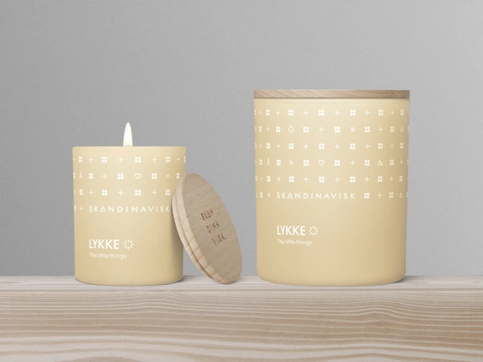 To gule lys i krukker med trælåg, det ene tændt, på en træoverflade. Begge krukker er mærket LYKKE, SKANDINAVISK og dekoreret med små hvide symboler.