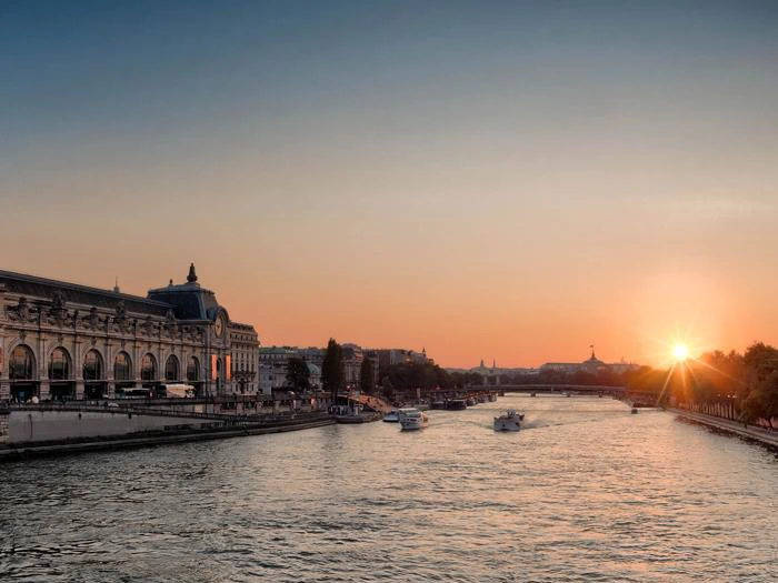 Solnedgang over Seinen i Paris med både på vandet og historiske bygninger, bl.a. Musée dOrsay, langs flodbredden.