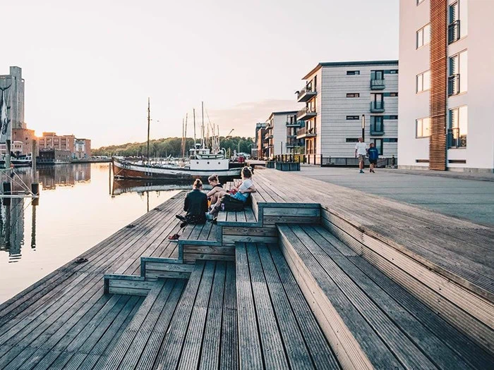 Folk sidder på trætrapper ved en havnefront med oplagte både og moderne bygninger ved solnedgang, hvilket skaber en rolig og afslappet atmosfære.
