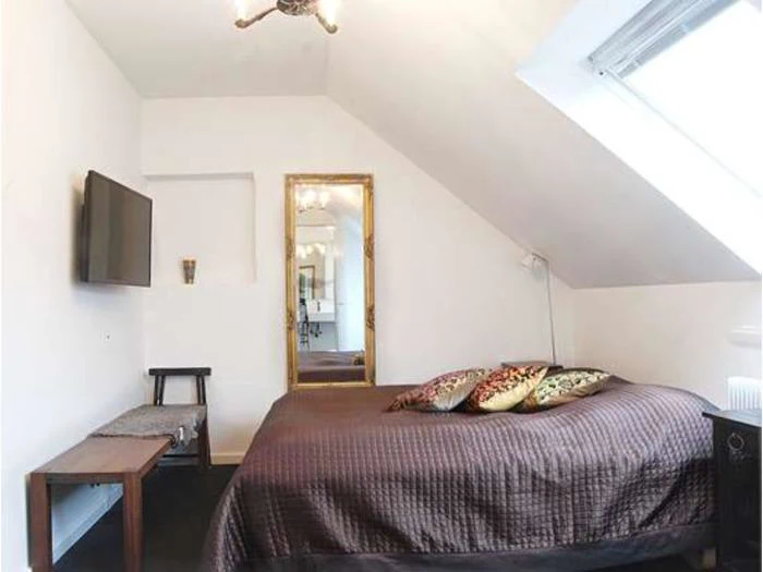 Soveværelse på loftet med skråt loft, brunt sengetæppe, pyntepuder, væghængt tv, bænk og et stort guldindrammet spejl, der reflekterer et andet rum.