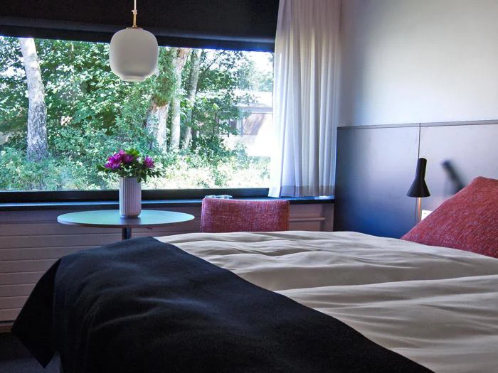 Et hyggeligt soveværelse med en seng, røde puder, et lille rundt bord med en vase med blomster, et vindue med udsigt til grønne træer og moderne belysning.