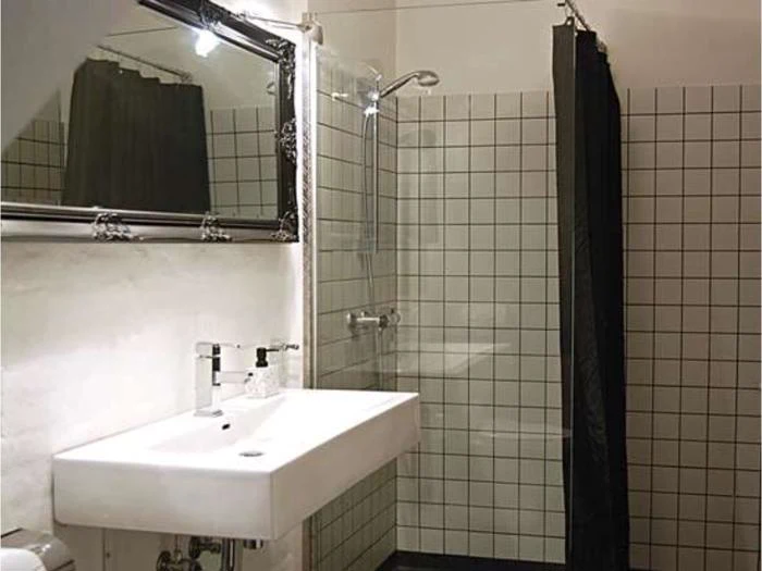Moderne badeværelse med rektangulær vask, væghængt vandhane, stort spejl og en brusekabine med hvide, kvadratiske fliser og et sort bruseforhæng.