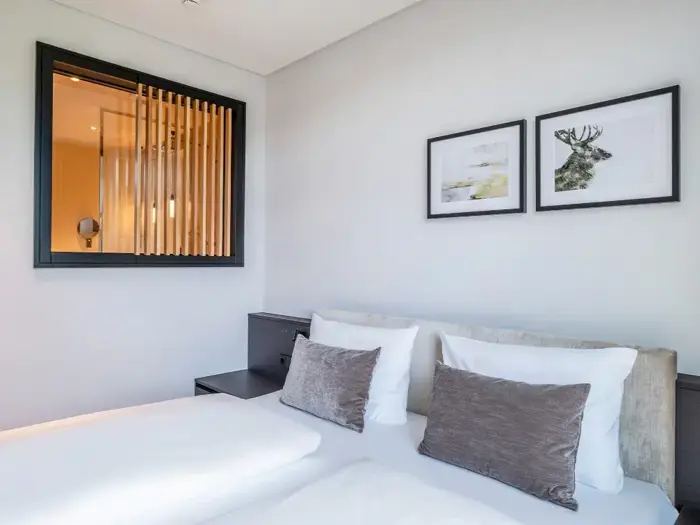 Et moderne soveværelse med en pænt redt seng, grå puder, to indrammede billeder på en hvid væg og et vindue med trælameller ind til et andet rum.