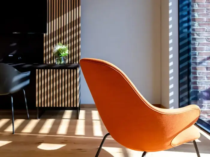 Et moderne rum med en orange lænestol, sollys, der strømmer ind gennem persienner og kaster skygger på trægulvet og væggen; et bord med en vase med blomster står i baggrunden.