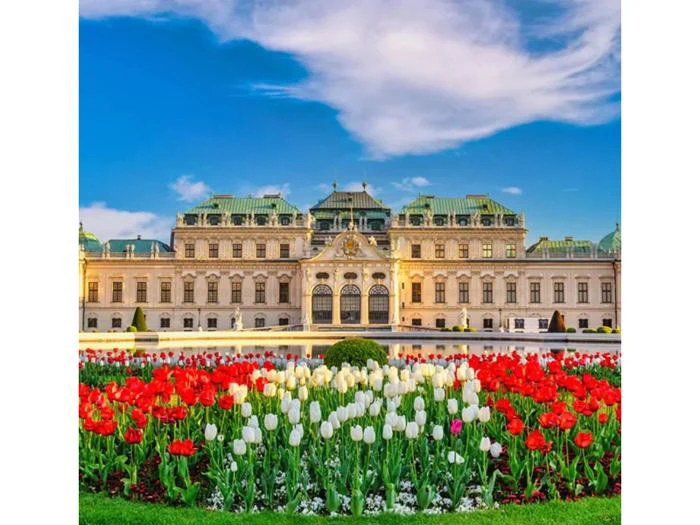 Et storslået palads med grønt tag står bag levende røde og hvide tulipaner i en velplejet have under en lyseblå himmel med tynde skyer.
