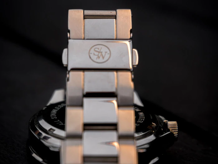 Nærbillede af et metalarmbånd med S/W-logoet indgraveret på låsen, og en del af urkassen er synlig i baggrunden.