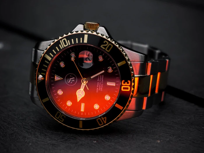 Nærbillede af et sølvfarvet og sort armbåndsur med gulddetaljer, oplyst af et orange lys, der hviler på en mørk overflade.