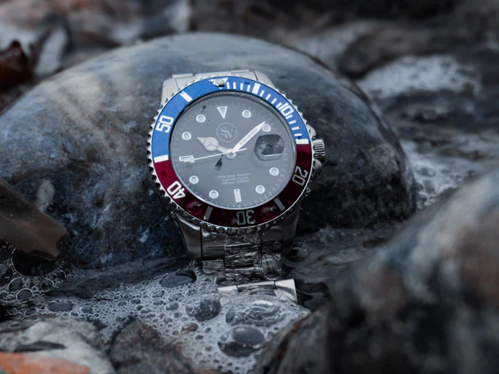 Et sølvfarvet armbåndsur med en blå og rød ramme hviler på en våd sten på lavt vand, omgivet af bobler og glatte sten.