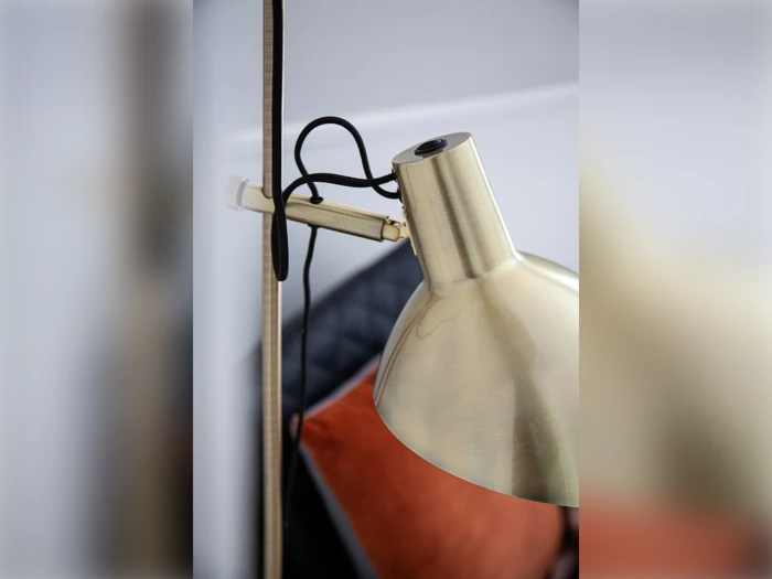 Nærbillede af en moderne skrivebordslampe i messing med sort ledning, placeret ved siden af en sort stol og en orange pude i baggrunden.
