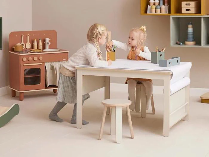 To små børn leger og tegner ved et hvidt bord i et pænt, pastelfarvet legeværelse med et legetøjskøkken og hylder i baggrunden.