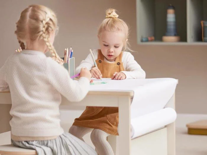 To små børn sidder ved et bord og tegner med farveblyanter på hvidt papir. Den ene har en brun kjole på, den anden en lys sweater og nederdel. Baggrunden består af hylder med dekorationer.