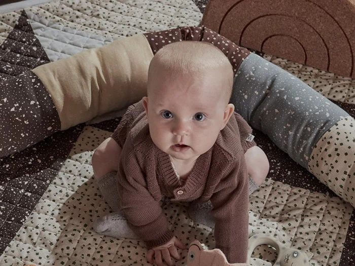 En baby iført en brun trøje og grå sokker sidder på et mønstret tæppe, omgivet af en blød, flerfarvet pude.