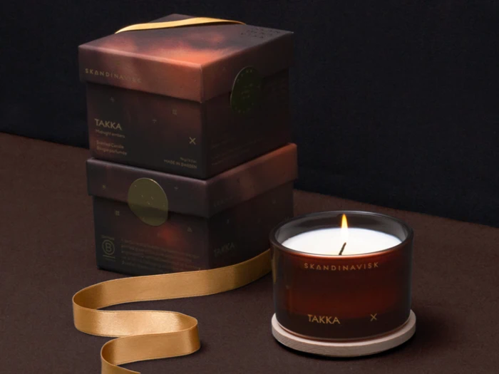 Et tændt stearinlys i en brun glasholder mærket TAKKA står på en bordskåner ved siden af to stablede matchende æsker og et guldbånd på en mørk overflade.