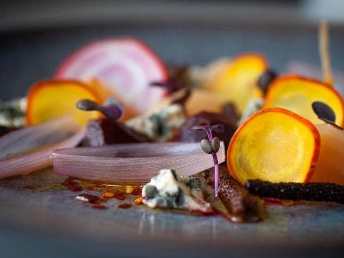 Et nærbillede af en kunstnerisk gourmetret med gyldne og røde rødbeder i skiver, syltede løg, mikrogrønt og smuldret blåskimmelost på en tallerken.
