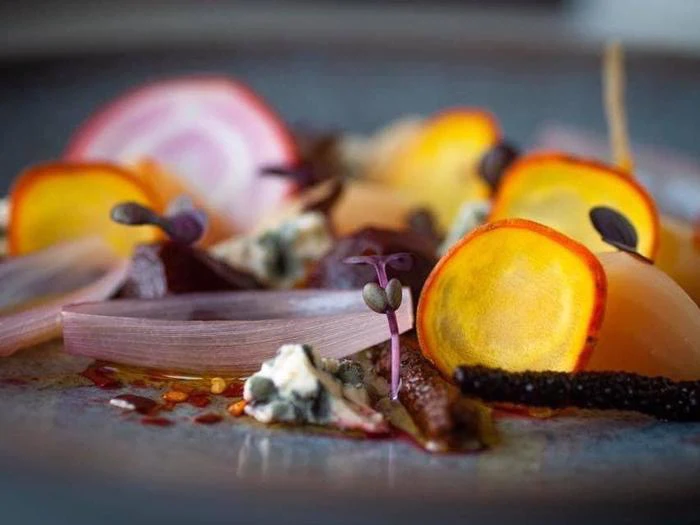 Et nærbillede af en gourmetret med tyndtskårne farverige grøntsager, mikrogrønt, ostecrumbles og kunstnerisk anretning på en grå tallerken.