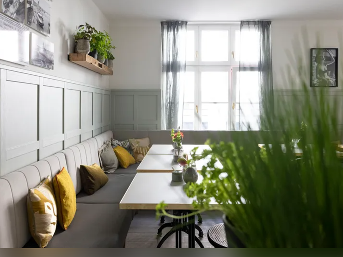 En lys og hyggelig café med hvide borde, bænke med hynder, gule puder, planter og sollys, der strømmer ind gennem et stort vindue med tynde gardiner.