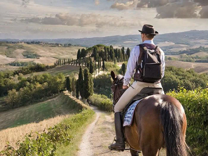 En person med hat og rygsæk rider på en hest langs en landlig sti med cypresser og udsigt over bølgende bakker under en overskyet himmel.