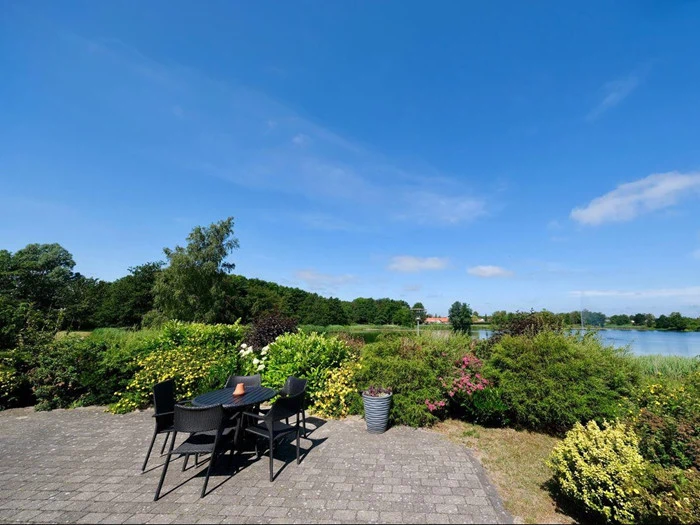 En terrasse med et sort bord og stole har udsigt til en frodig have, en sø og træer under en klar blå himmel.