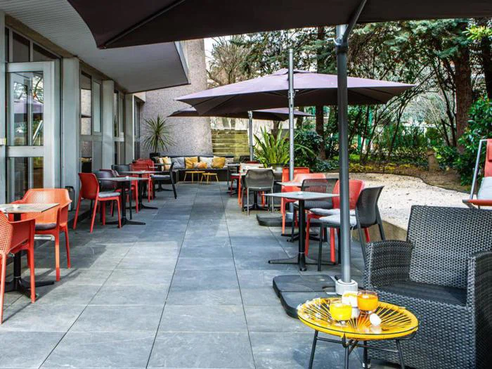 Udendørs gårdhave ved en café med forskellige stole og borde, skygget af store parasoller, omgivet af planter og grønt. Drikkevarer og kager står på et gult bord i forgrunden.