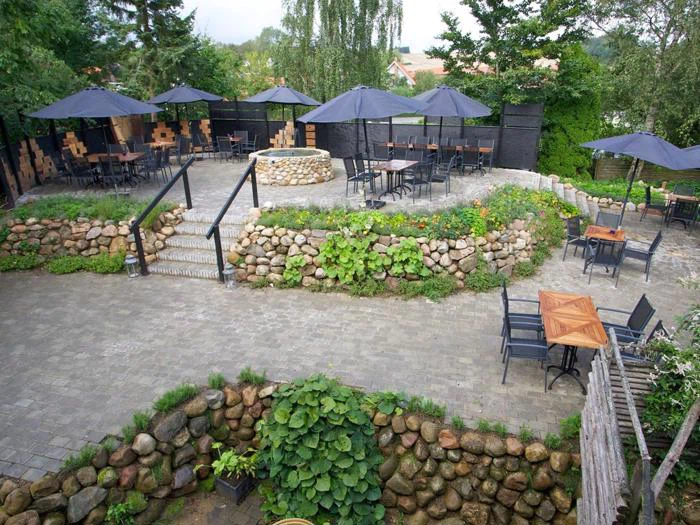 Udendørs gårdhave med stenvægge, borde og stole, omgivet af grønne planter og sorte parasoller, fordelt på forskellige terrasseniveauer med en cirkulær sten i midten.