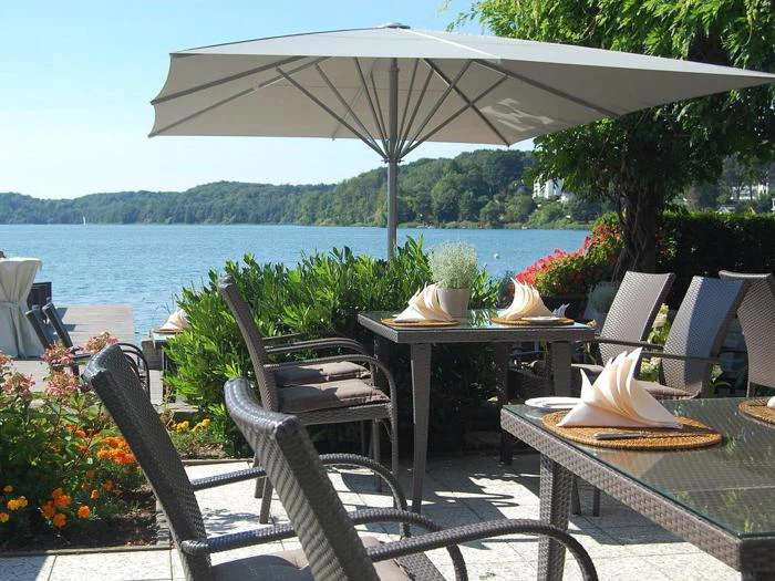 Udendørs spiseplads med kurvestole, dækkede borde med foldede servietter, stor parasol, blomster og en naturskøn udsigt over søen med grønne bakker i baggrunden.
