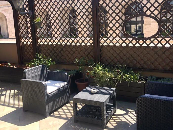 Udendørs terrasse med sorte kurvemøbler, potteplanter og trægitterpaneler, der kaster skygger, med et rundt spejl på væggen, der reflekterer en del af bygningen.