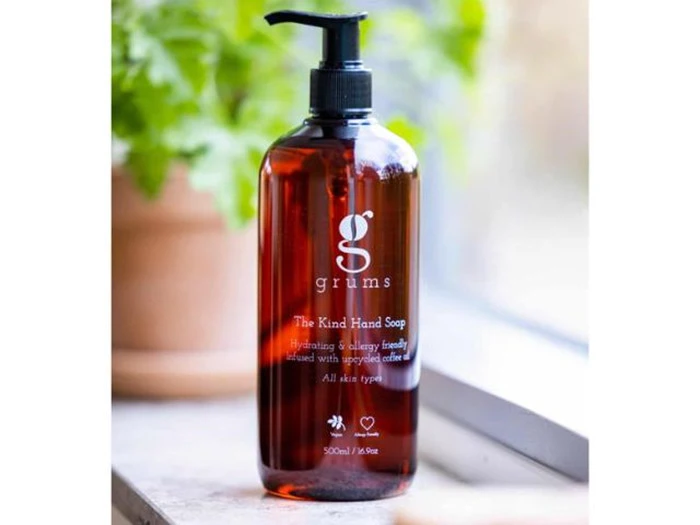 En brun pumpeflaske med Grums The Kind Hand Soap står i en vindueskarm med en grøn plante sløret i baggrunden.