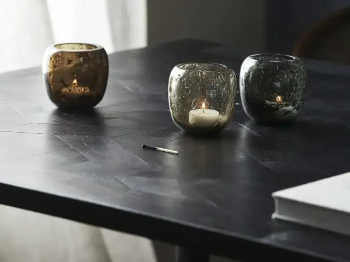 Tre glaslysestager med tændte fyrfadslys står på et mørkt bord ved siden af en bog og en brugt tændstik. Blødt naturligt lys kommer fra et vindue i baggrunden.